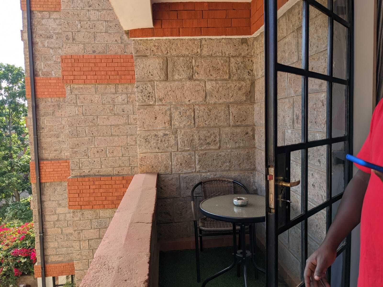 Un balcón con pared de ladrillo, una mesita con una silla y una puerta con reja metálica.