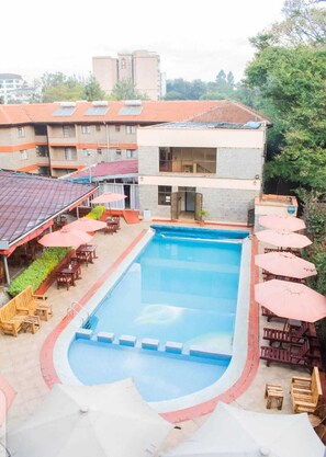 A swimming pool with surrounding wooden furniture and umbrellas, a building with a tiled roof, and greenery in the background. - Bliss haven apartments  (Nairobi)