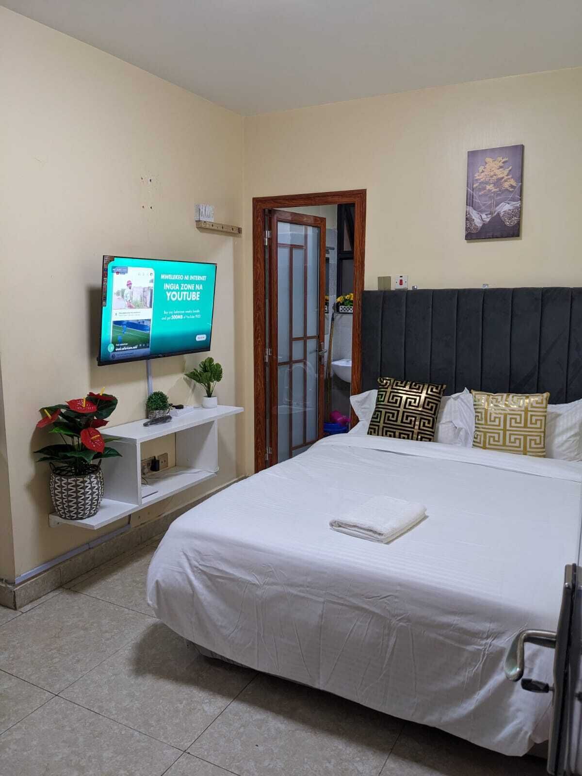 A bedroom with a bed, a TV on the wall, a door, and a shelf with plants.