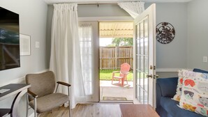 A room with a wooden floor, a blue sofa, a chair, a small table, and a door leading to an outdoor area with a chair and a wooden fence.
