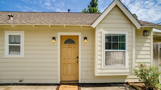 A single-story house with a beige exterior, a wooden front door, two windows, and a small porch area.