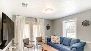 A living room with a blue sofa, two chairs, a wooden table, a flat-screen TV, a clock, and a window with curtains.