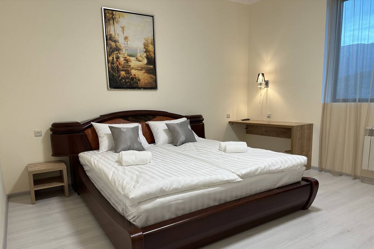 Una cama bien tendida con sábanas y almohadas blancas, una pequeña mesita de noche, una lámpara de pared y un cuadro sobre la cama.