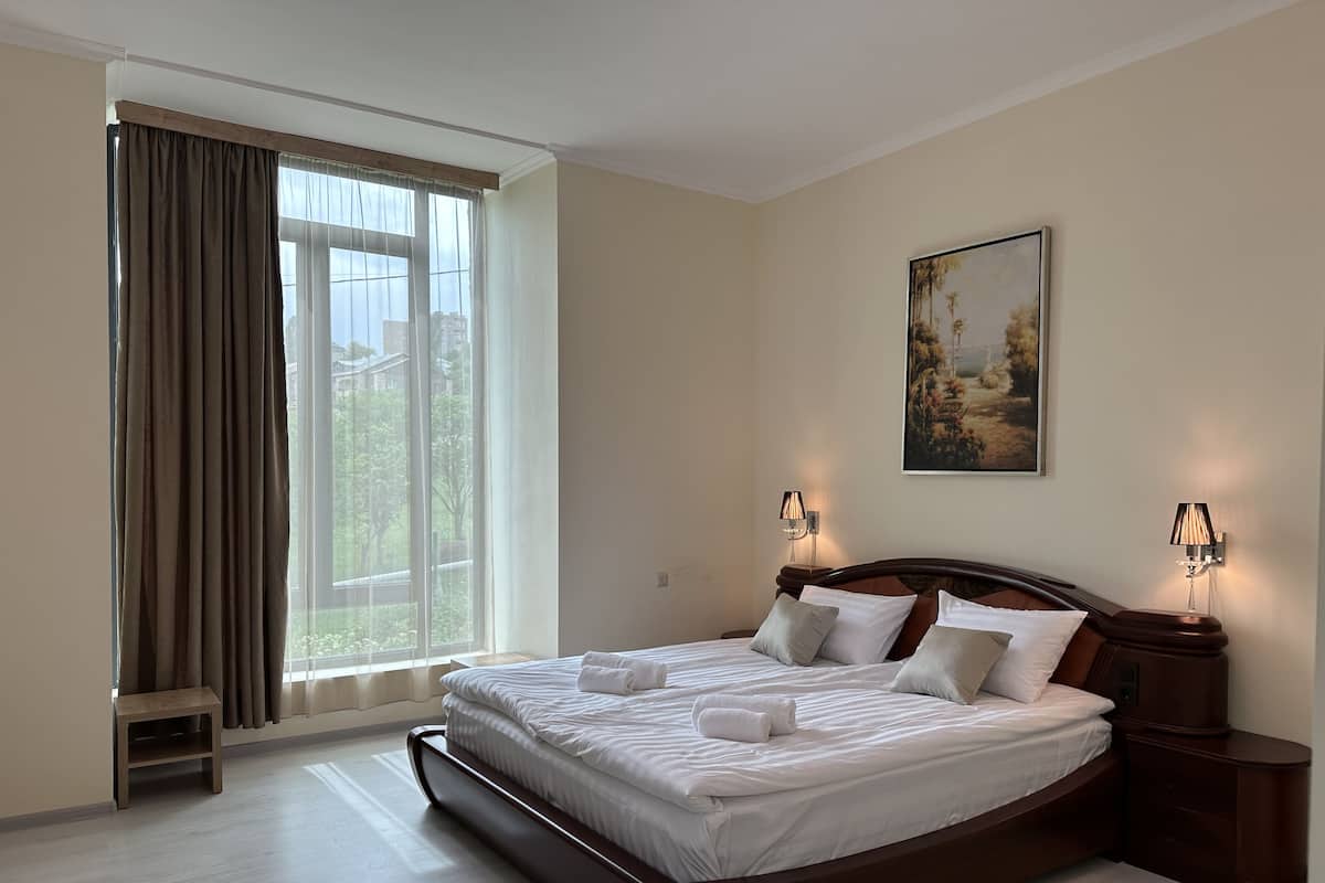 Una cama bien hecha con sábanas y almohadas blancas, una mesita de noche con lámpara, un cuadro en la pared y una ventana con cortinas.