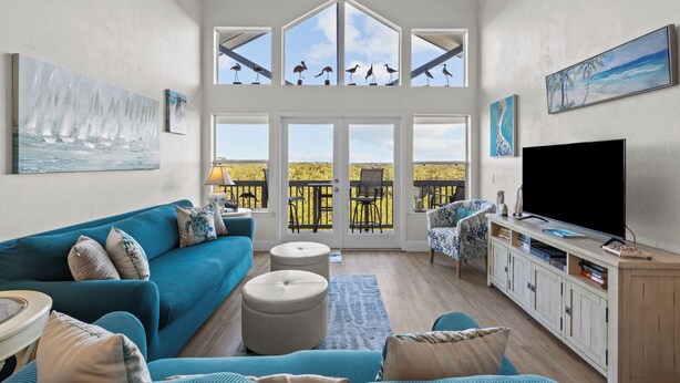 A spacious living room with a large window, a blue sofa, a TV, and a wooden cabinet. - Fenimore Mill 10E - Pool, Hot Tub, Boat Parking! (Cedar Key)