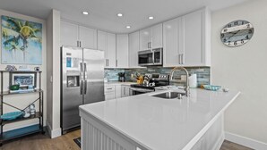 A modern kitchen with white cabinetry, stainless steel appliances, a large island with a sink, and a clock on the wall. - Fenimore Mill 10E - Pool, Hot Tub, Boat Parking! (Cedar Key)