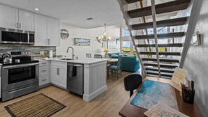 A modern kitchen with white cabinetry, a built-in microwave, and a stove. There is a dining area with a blue sofa and a staircase leading to a loft. - Fenimore Mill 10E - Pool, Hot Tub, Boat Parking! (Cedar Key)