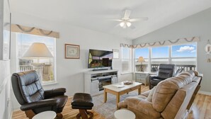 Ein Wohnzimmer mit Fernseher, Ledersesseln, einer Couch, einem Deckenventilator und großen Fenstern.