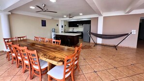 A spacious dining area with a long wooden table and matching chairs, a kitchen in the background, and a hammock against the wall. - Quinta del Carmen (La Libertad)