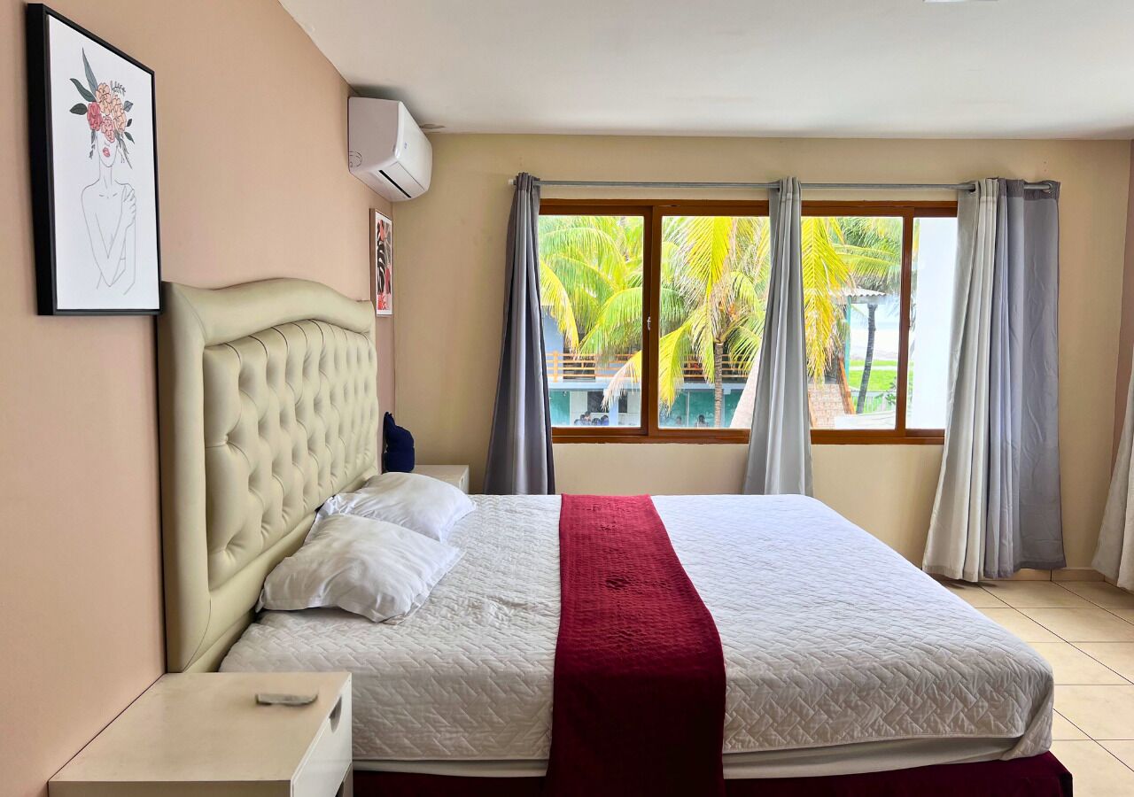 Ein Schlafzimmer mit einem großen Bett, einem Kopfteil, einem Nachttisch, einem Fenster mit Vorhängen und Blick auf Palmen im Freien.