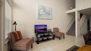 A living room with a flat-screen TV, patterned sofas, a staircase, and a painting on the wall. - Fenimore Mill 2E - Open Gulf, Pool, Hot Tub! (Cedar Key)