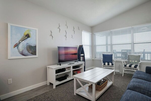 A living room with a flat-screen TV, white entertainment unit, and a coffee table. There are two white chairs with striped cushions, a blue sofa, and a large window with blinds. - Fenimore Mill 2F - Gulf Front, Pool, Pet Friendly! (Cedar Key)