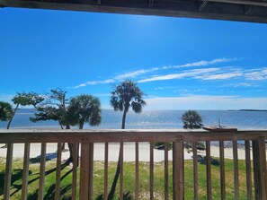 A wooden deck overlooking a beach with palm trees and a clear blue sky. - Fenimore Mill 4B - Pool, Hot Tub, Open Gulf View! (Cedar Key)