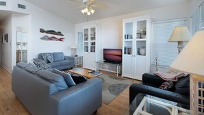 A modern living room with a grey sofa, a black sofa, a glass coffee table, a TV, and a ceiling fan. - Fenimore Mill 4F - Open Gulf View, Pool, Hot Tub! (Cedar Key)