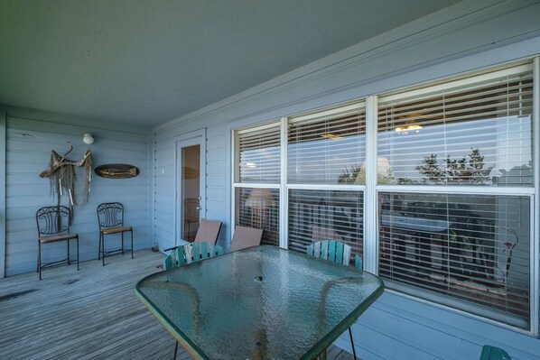 A covered porch with a glass table and metal chairs, wooden walls, and a large window with blinds. - Fenimore Mill 4F - Open Gulf View, Pool, Hot Tub! (Cedar Key)