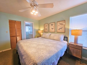 A bedroom with a bed, a ceiling fan, a nightstand with a lamp, and framed artwork on the wall.