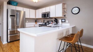 A modern kitchen with white cabinetry, stainless steel appliances, and a central island with bar stools. - Fenimore Mill 7A - Beautiful View, Pool, Hot Tub! (Cedar Key)