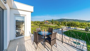 Een balkon met een tafel en stoelen, een glazen deur en uitzicht op een groen landschap met heuvels en een huis in de verte.