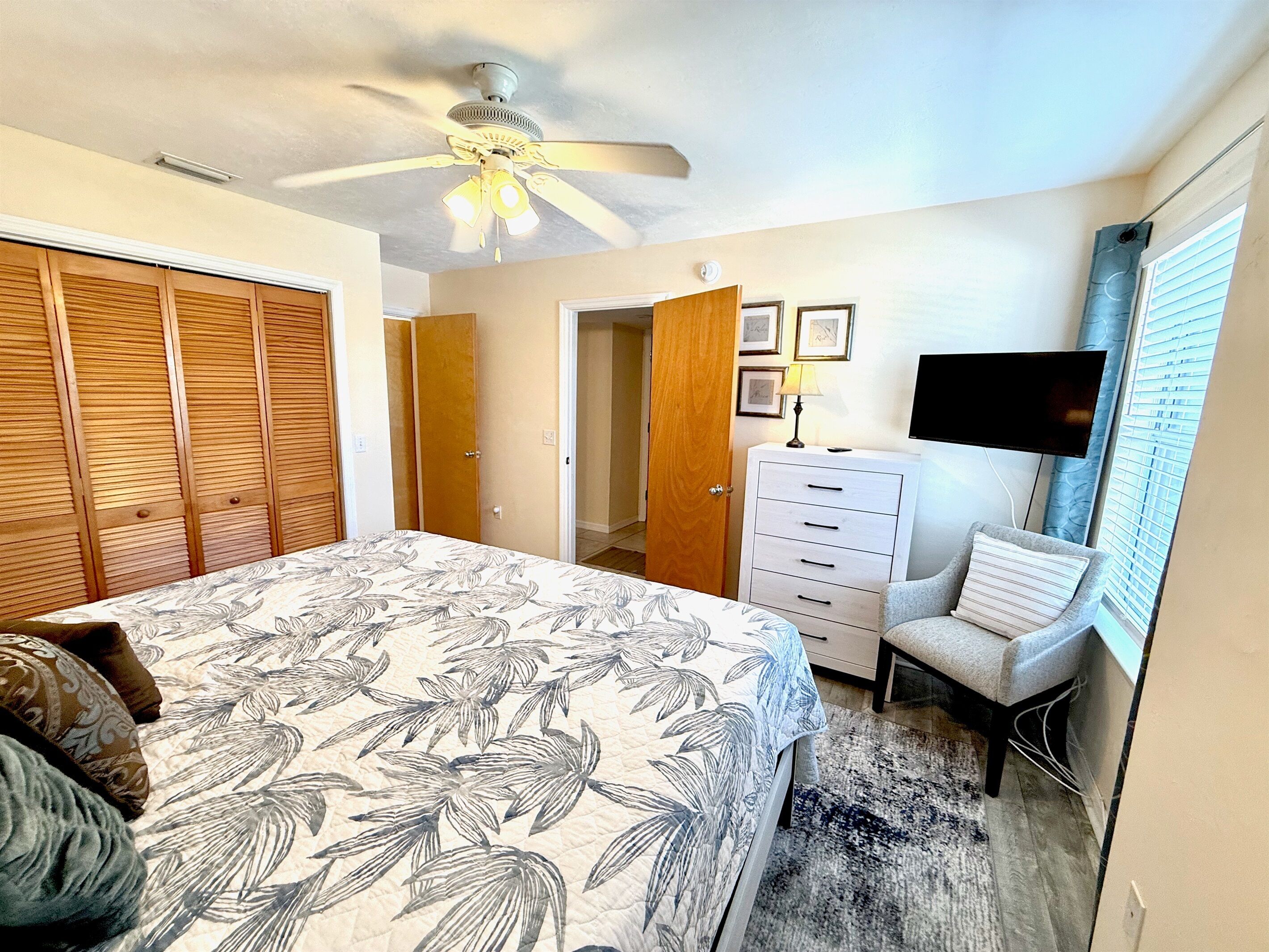 A bedroom with a bed, a ceiling fan, a dresser, a chair, a TV, and a door leading to another room.