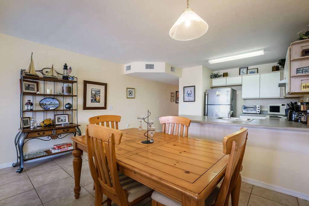 A dining area with a wooden table and chairs, a chandelier, and a kitchen in the background.