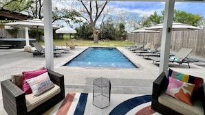 Un área de piscina exterior techada con sillas de descanso y sombrillas, rodeada de árboles y una valla de madera.