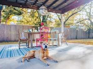 A dog lying on a patio with a covered table and chairs, a hanging light fixture, and a wooden fence in the background.