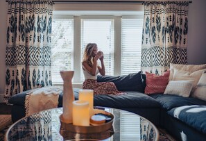 A cozy living room with a large window, patterned curtains, a sofa with cushions, and a coffee table with candles.