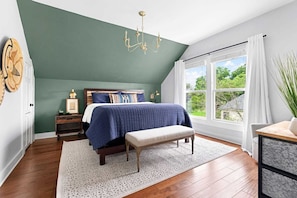A bedroom with a sloped ceiling, a large window with curtains, a bed with a blue blanket, a wooden bench, and a chandelier. - Fredericksburg 3BR · Pool, Hot Tub · Sleeps 8 (Fredericksburg)