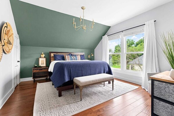 A bedroom with a sloped ceiling, a large window with curtains, a bed with a blue blanket, a wooden bench, and a chandelier.