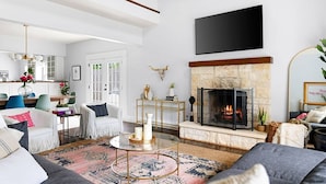 Una sala de estar moderna con chimenea, un televisor de pantalla plana montado en la pared y un comedor con mesa y sillas blancas.