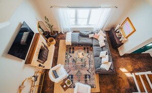 Ein geräumiges Wohnzimmer mit großem Fenster, einem grauen Sofa, einem gemusterten Teppich und einem Fernseher an der Wand.