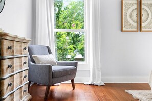 Une pièce avec un fauteuil gris, une commode en bois et une fenêtre ornée de rideaux blancs.