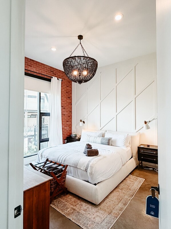A modern bedroom with a large bed, a hanging light fixture, a brick accent wall, and a view of the city.