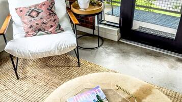 A cozy room with a white chair, a round table with a magazine and two cups of green tea, and a view of greenery outside.