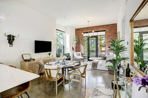 Een moderne woonkamer met een accentmuur van baksteen, een ronde eettafel met stoelen, een flatscreen televisie aan de muur bevestigd en een groot raam dat natuurlijk licht biedt.