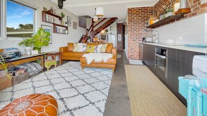 En hyggelig stue med sofa, sofabord og bogreol. Der er et køkkenområde med komfur og vask.