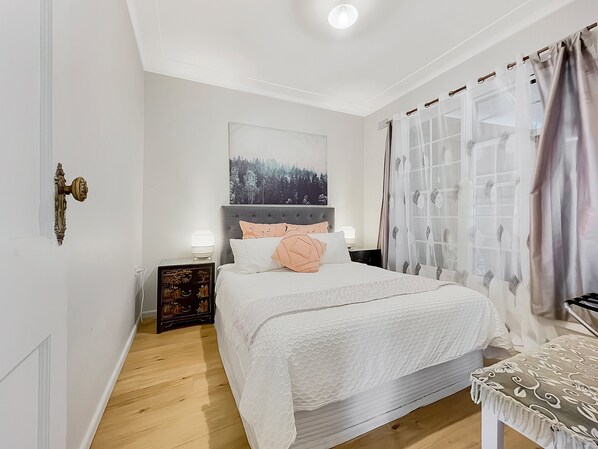 A bedroom with a bed, a nightstand, a bedside table, a window with curtains, and a door with a handle. - Sea Dreaming - Pambula Beach (Pambula Beach)