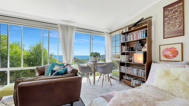 Une pièce spacieuse dotée d’une grande fenêtre donnant sur la mer, un coin salon confortable avec un canapé marron et un lit moelleux recouvert d’une couette blanche, ainsi qu’une bibliothèque remplie de livres et d’objets décoratifs.