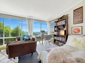 A spacious room with a large window offering a view of the sea, a cozy seating area with a brown sofa and a fluffy white bed, and a bookshelf filled with books and decorative items. - Sea Dreaming - Pambula Beach (Pambula Beach)
