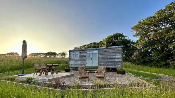 A rustic cabin with a wooden exterior, a small porch with outdoor seating, and a clear view of the surrounding grassy landscape.