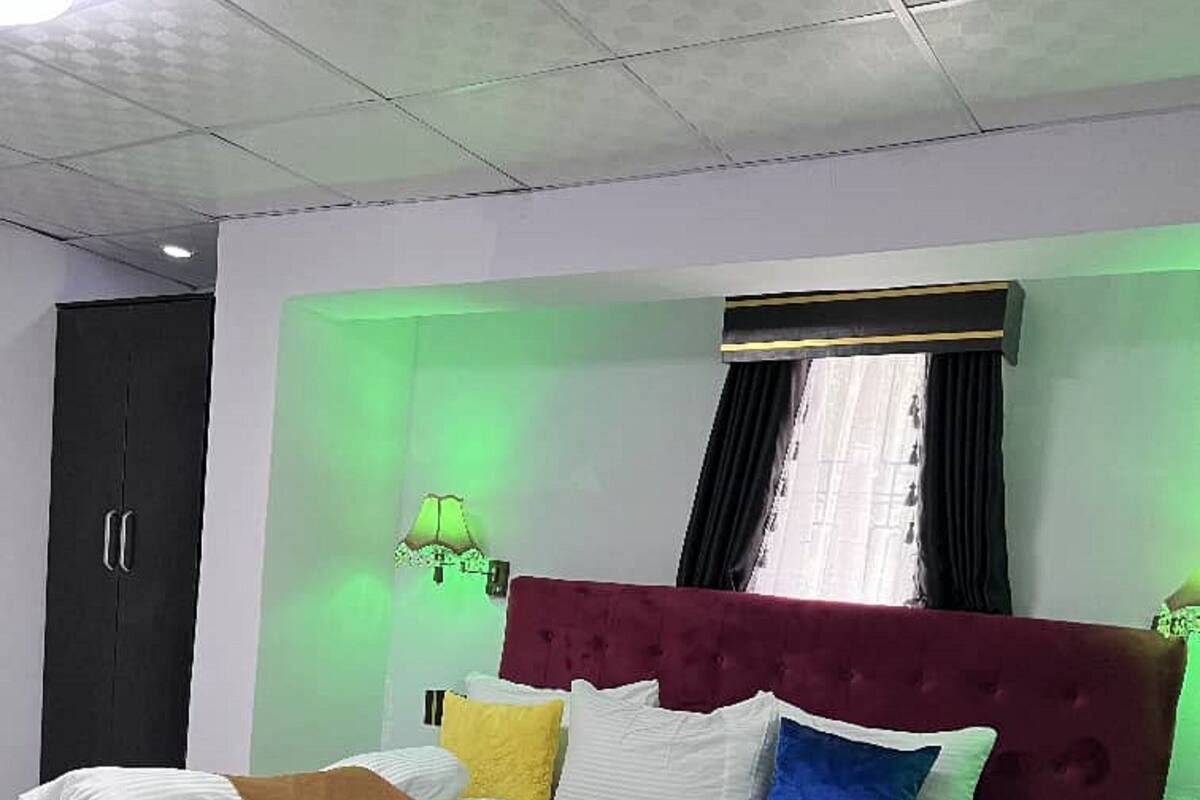 Una cama bien hecha con cabecera roja, una mesita de noche con lámpara y un armario en una habitación con iluminación verde ambiental.