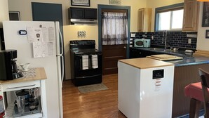 Une cuisine compacte équipée d’un réfrigérateur, d’une cuisinière, d’un four à micro-ondes et d’un petit évier. On y trouve des armoires en bois, une machine à café et un tapis sur le sol.