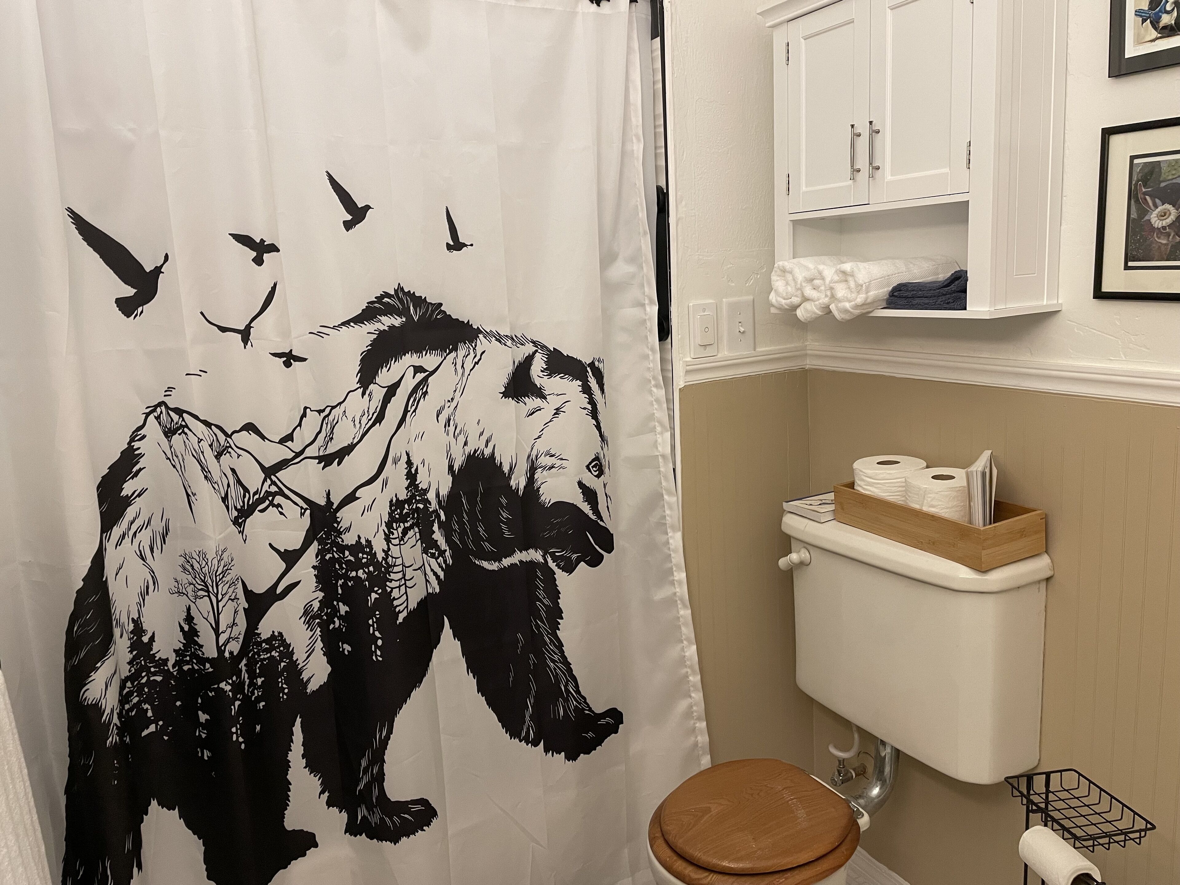 Et badeværelse med bruseforhæng med et bjergmotiv, et toilet med træsæde og en hylde med toiletartikler.
