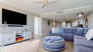 Ein modernes Wohnzimmer mit Flachbildfernseher, Deckenventilator, grauem Ecksofa, rundem Pouf und einer Küche im Hintergrund.
