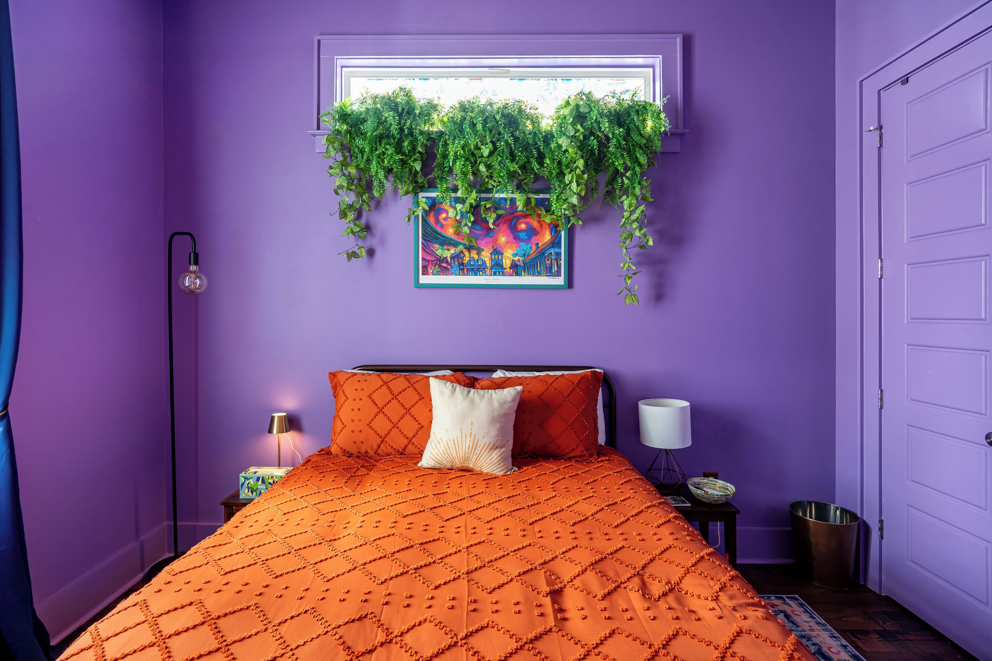 Une pièce avec un lit, une fenêtre ornée de plantes, un tableau et une porte.