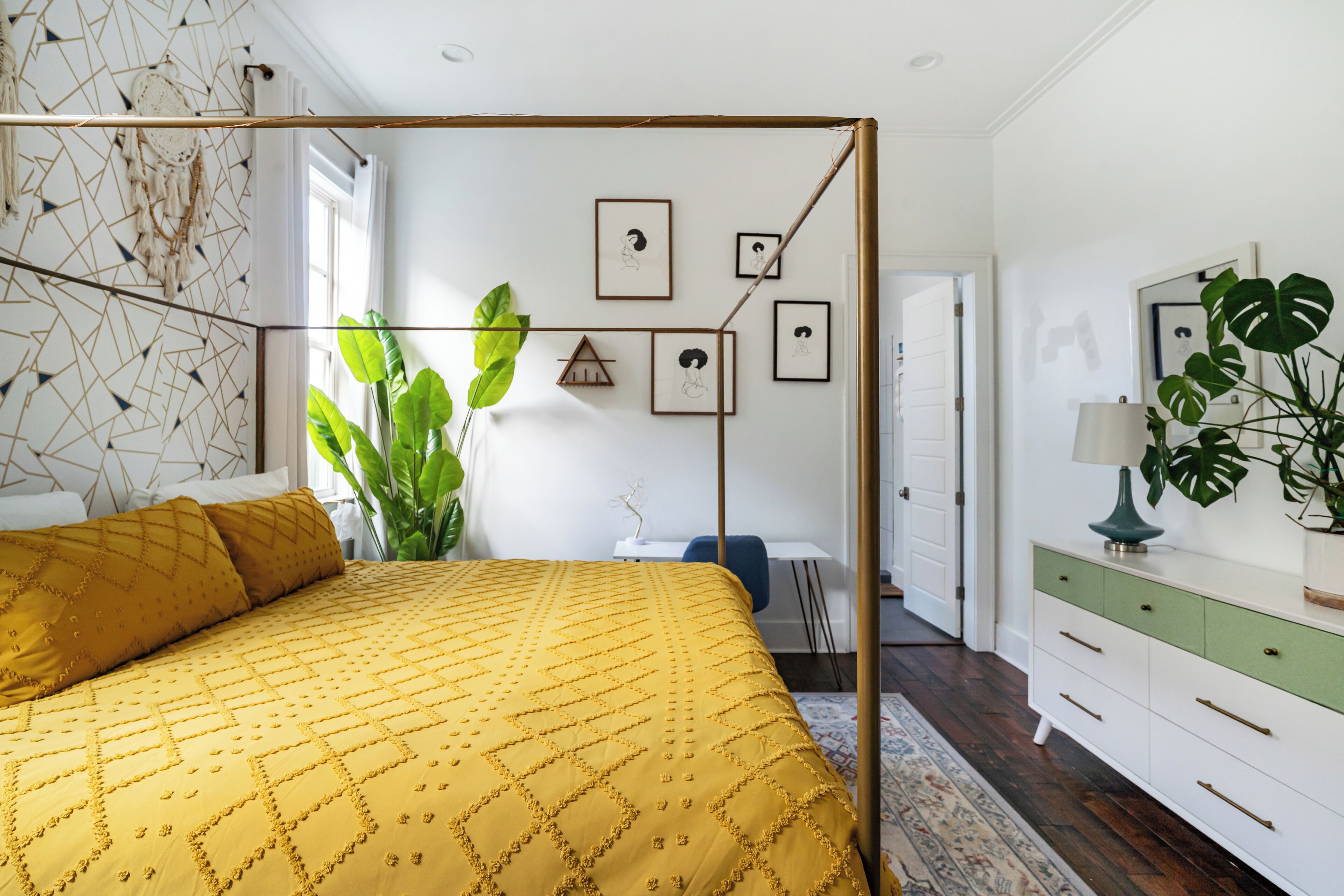Ein Schlafzimmer mit Himmelbett, einer großen Grünpflanze, einem Schreibtisch und einer Kommode.