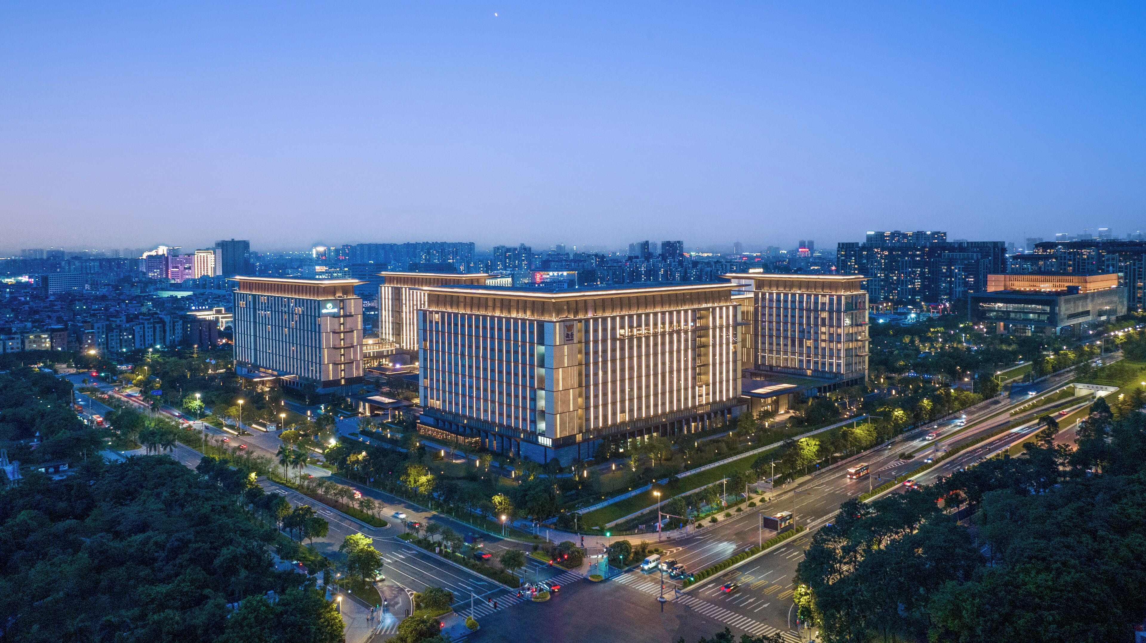 Un immeuble moderne de grande hauteur, de forme rectangulaire, entouré d’un environnement urbain bien aménagé, avec des rues et des espaces verts.