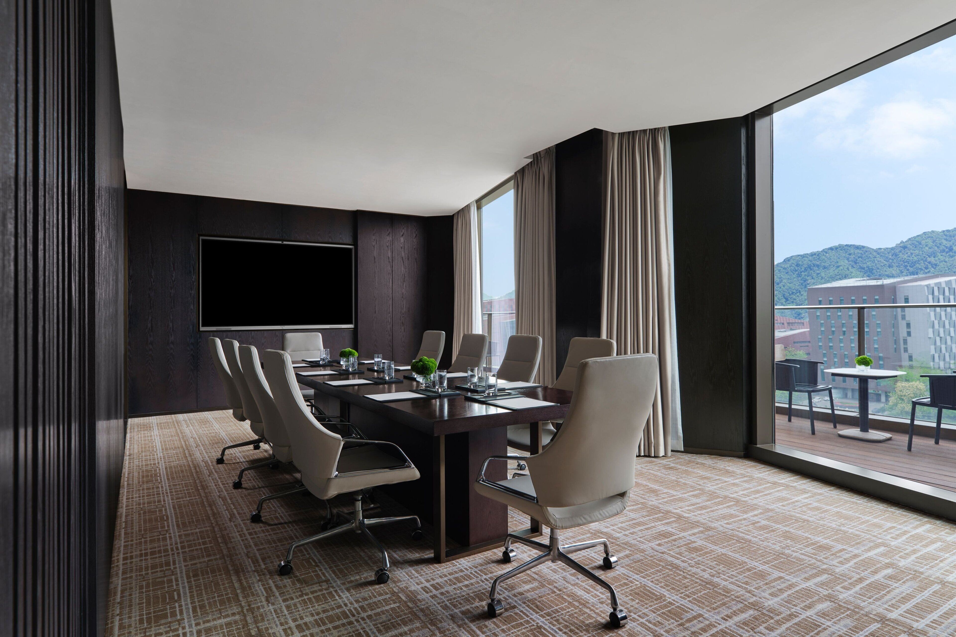 A conference room with a long table, multiple chairs, a flat-screen TV, and large windows offering a view of a cityscape and mountains.
