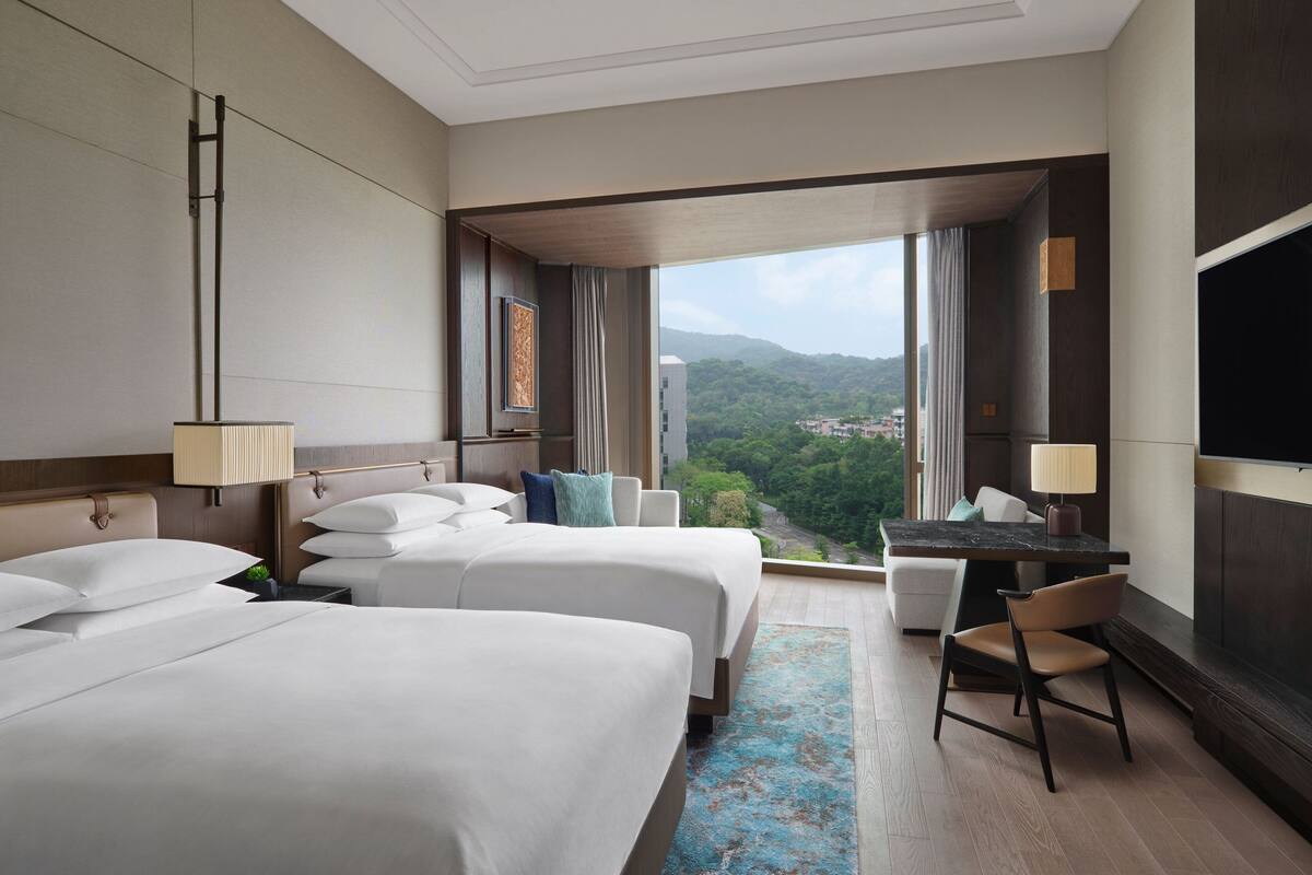Une chambre d’hôtel avec deux lits, un bureau, une chaise et une grande télévision. La chambre offre une vue sur la ville et les montagnes.