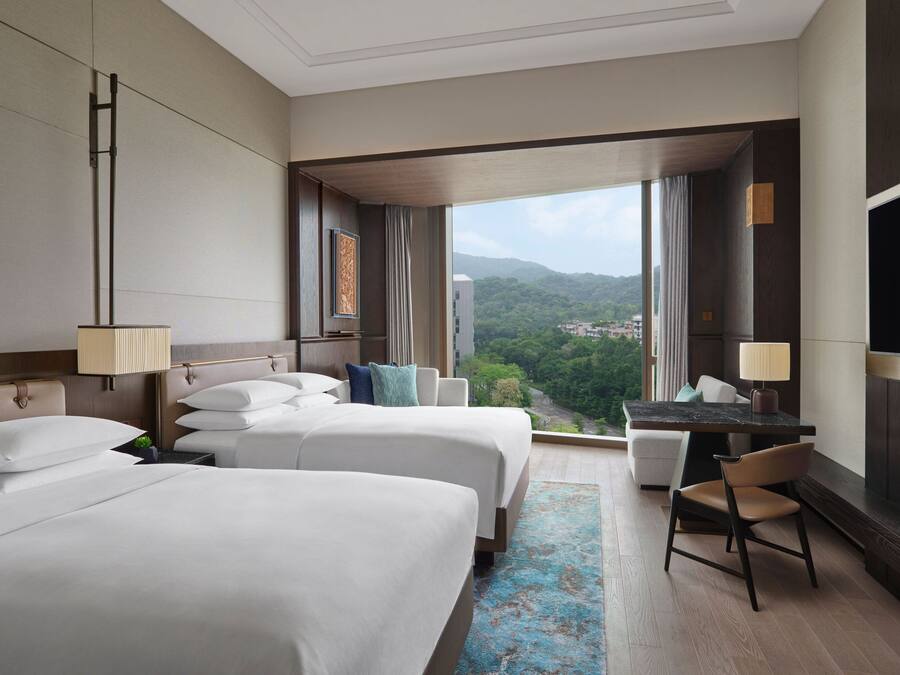 A hotel room with two beds, a desk, a chair, and a large TV. The room has a view of a city and mountains.